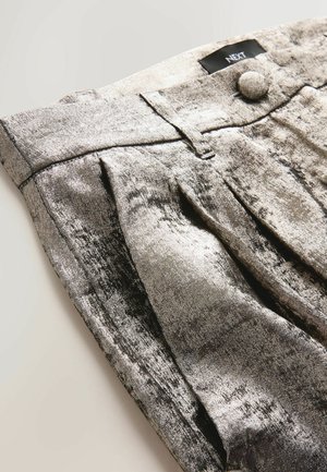 Pantaloni plisati metalici argintii cu textură moale, având detalii la nasturele din față și la talie, expuși dintr-un unghi. Eticheta brandului este vizibilă.