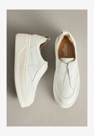 Hvide slip-on sko i læder med rund tå, tekstureret overflade, kontrasterende syninger og en hvid gummisål. Har en trækkefane ved hælen.