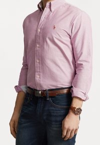 Jasnoróżowa koszula zapinana na guziki z długimi rękawami, z małym logo na piersi. Noszona z ciemnoniebieskimi dżinsami i brązowym skórzanym zegarkiem.