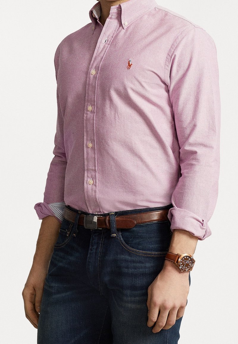 Jasnoróżowa koszula zapinana na guziki z długimi rękawami, z małym logo na piersi. Noszona z ciemnoniebieskimi dżinsami i brązowym skórzanym zegarkiem.