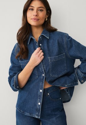 Kvinde med langt brunt hår iført en oversize mørk denimjakke og matchende jeans, justerer jakken foran med den ene hånd.