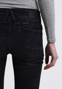Jeans de mezclilla negra con corte ajustado, que cuentan con bolsillos traseros, costura visible y una superficie texturizada. La cinturilla tiene una etiqueta de cuero.
