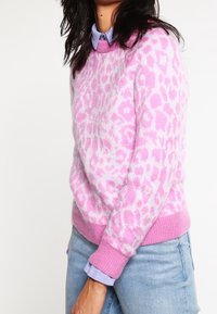 Pull en imprimé léopard rose avec un col et des poignets côtelés courts. Superposé sur une chemise à col bleu clair. Tissu en maille doux et texturé.