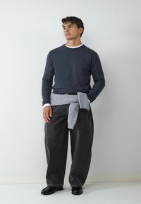 Donker langlopende shirt met witte kraag, grijze trui om de taille geknoopt, ruime donkere jeans en zwarte schoenen. Eenvoudig, modern design.
