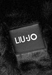 Materiale in pelliccia sintetica nera con una patella in pelle liscia che presenta il testo bianco "LIU·JO". La texture è morbida con un leggero lucido.