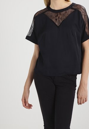 Vrouw die een zwart topje met korte mouwen draagt, voorzien van doorschijnende kanten panelen op de schouders en de bovenkant van de borst, gecombineerd met zwarte strakke broek.