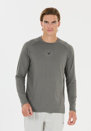 Mand iført en grå træningsshirt med lange ærmer og lyse bukser, stående foran en ensfarvet hvid baggrund.