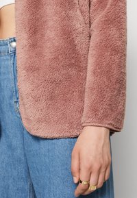 Fuzzy rosa jacka med mjuk textur, rundad nederkant och långa ärmar, ihop med högmidjade blå jeans. Innehåller guldringar på fingrarna.