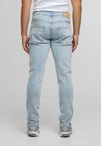 A kép hátulról mutat egy személyt, aki világoskék, slim-fit farmernadrágot, fehér topot és ezüstszürke tornacipőt visel, egy sima szürke háttér előtt.