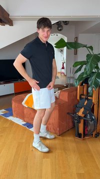 Young man in black shirt and white shorts standing in a living room with wooden floor, orange sofa, large green plant, and rowing machine.