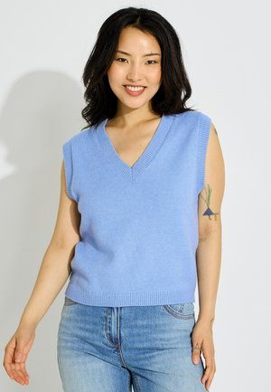 Joven sonriente, lleva un chaleco de punto azul claro sin mangas y vaqueros azul claro, con un pequeño tatuaje en su brazo izquierdo superior.