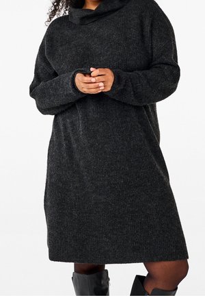 Kvinde iført mørkegrå, langsleeve strikket kjole og sorte knæhøje støvler, stående med hænderne foldet foran livet.