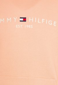 Sudadera color durazno con logotipo bordado "TOMMY HILFIGER" y símbolo de bandera. Textura suave, cuello redondo y corte relajado.