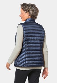 Marineblauer Pufferweste mit hohem Kragen, gestepptem Design und leichtem Glanz, kombiniert mit einem cremefarbenen, strukturierten Pullover darunter.