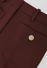 Pantalones de tela burdeos con un solo bolsillo frontal, que cuentan con un botón circular dorado y costuras reforzadas. Diseño liso y entallado.