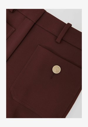 Bordeauxfarvede bukser i stof med en enkelt lomme foran, der har en rund guldknap og forstærkede sømme. Glat, skræddersyet design.