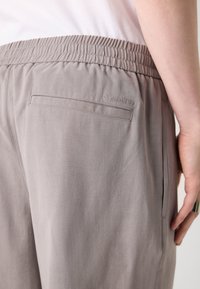 Gros plan sur un pantalon beige à taille élastique avec une poche arrière et un logo discret Calvin Klein au-dessus de la poche, sur la hanche d’une personne.