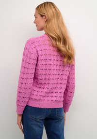 Maglioncino rosa lavorato a maglia con un motivo in pizzo testurizzato, polsini e orlo a costine. Abbinato a jeans blu.