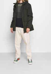 Giacca con cappuccio verde scuro con due tasche frontali, indossata su un maglione a righe blu navy e verde, abbinata a pantaloni beige e sneaker multicolori.