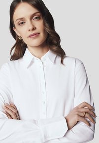 Biała koszula z długim rękawem, o fakturowanej tkaninie i klasycznym kołnierzyku, z rzędem guzików z przodu.