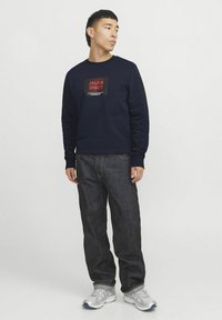 Marineblauwe sweatshirt met een rechthoekig rood en zwart logo op de borst, gecombineerd met donkere denim jeans en lichte sneakers.