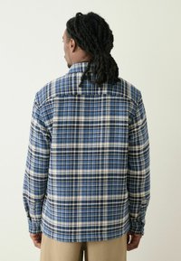 Plaid shirt in blue, white, and navy featuring a classic collar, long sleeves, and a textured fabric. Back view shows the pattern clearly.