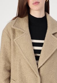 Béžový texturovaný kabát s klopovým límcem, doplněný černým žebrovaným svetrem s vodorovnými bílými pruhy.