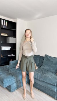 Beige langemouwen top, olijfgroene shorts met een grote ronde ceintuurgesp, en nude hoge hakken. Achtergrond met een donkere boekenkast.