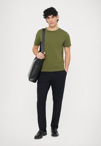 Olivově zelené tričko, úzký střih, kombinované s tmavě modrými kalhotami. Černá kožená taška přes rameno, lesklé černé boty, studiové pozadí.