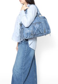 Borsa a tracolla blu in pelle con diverse tasche, dettagli in frange e hardware dorato. Indossata sulla spalla, abbinata a una camicia azzurro chiaro e jeans.