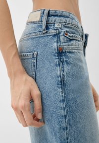 Primer plano de una persona que lleva puestos unos jeans de mezclilla azul claro con una mano descansando cerca del bolsillo trasero, frente a un fondo neutro.