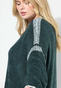 Donna bionda che indossa un maglione texturizzato verde scuro con motivi a spina di pesce bianchi e strisce sulle spalle e sulle braccia.