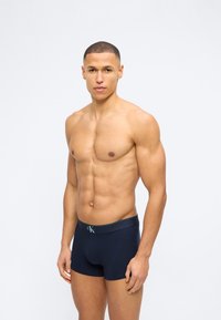 Αθλητικός άντρας με κοντά μαλλιά, στέκεται γυμνόστηθος, φοράει σκούρο μπλε μπόξερ με το λογότυπο Calvin Klein στη μέση.