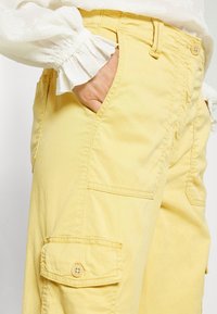 Hand in de zak van gele cargo-broek, gedragen met een witte blouse met lange mouwen en gerimpelde manchetten.