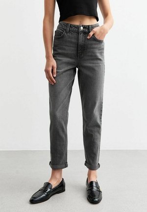 Osoba nosící ohrnuté tmavě šedé džíny s vysokým pasem, černý krátký top a lesklé černé mokasíny stojící na šedé podlaze u bílé zdi.