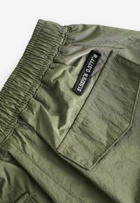 Pantaloni cargo verde oliva con cintura elastica, caratterizzati da una tasca applicata e un'etichetta nera che mostra le coordinate. Tessuto leggero e testurizzato.