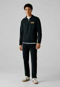 Młody mężczyzna w czarnej rozpinanej bluzie Red Bull Racing, czarnych spodniach i białych sneakersach stoi na tle jednolitego szarego tła.