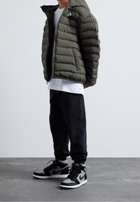 Olivengrüne Steppjacke mit Reißverschluss und Kapuze, weißes T-Shirt, schwarze Jogginghose und schwarze/graue High-Top-Sneaker mit Nike-Branding.