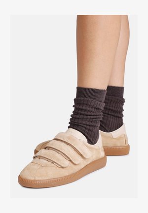 Bež osnovne športne čevlje iz usnja z dvema ježkoma, gumijastim podplatom in natančnim šivom, v kombinaciji z temno rebrastimi nogavicami do kolen.