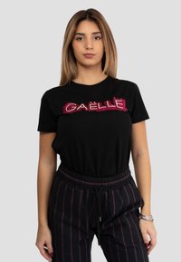 T-shirt nera con lettering "GAELLE" rosso testurizzato sulla parte anteriore. Maniche corte, collo rotondo, abbinata a pantaloni neri a righe.