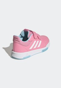 Zapatilla deportiva rosa con una parte superior sintética, forro azul, diseño de tres franjas blancas y una correa de velcro. Cuenta con una suela blanca con detalles en azul.