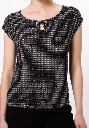Femme portant un haut à manches courtes à motif noir et blanc avec une petite ouverture devant en forme de goutte et un détail noué au niveau du col.