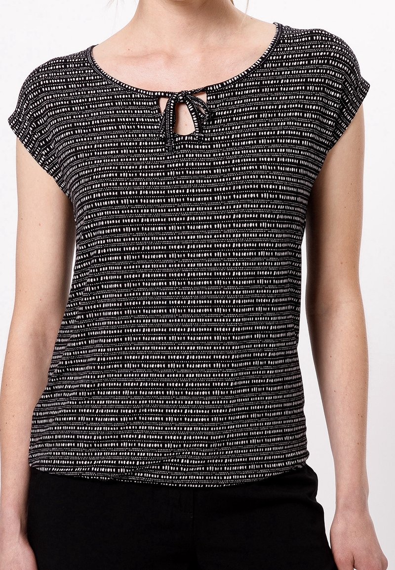 Femme portant un haut à manches courtes à motif noir et blanc avec une petite ouverture devant en forme de goutte et un détail noué au niveau du col.
