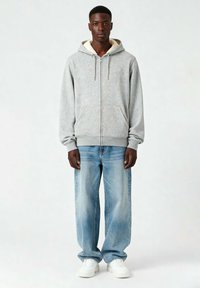 Felpa grigia con zip e fodera morbida color crema, tasche frontali e cordini, abbinata a jeans a gamba larga blu chiaro e sneakers bianche.