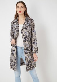 Trench coat con estampado floral en beige con acentos en azul y negro, que presenta un cuello de pico, cierre de botones y mangas tres cuartos.