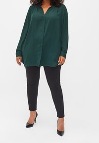 Blusa verde in satin con maniche lunghe, scollatura a V e taglio morbido, abbinata a pantaloni neri aderenti e tacchi alti neri.