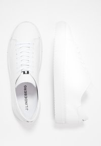 Bílé kožené tenisky s plochými tkaničkami, kulatou špičkou a texturovanou gumovou podrážkou. Mají logo na jazyku a minimalistický design.