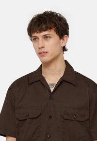 Brown short-sleeve shirt with a collar, two front pockets, and black buttons. Smooth texture and structured design for a casual look.