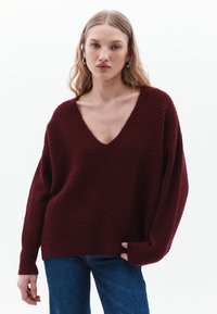 Bordeaux gebreide trui met een diepe V-hals, ribbelstructuur, verlaagde schouders en een losse pasvorm, gecombineerd met blauwe denim jeans.