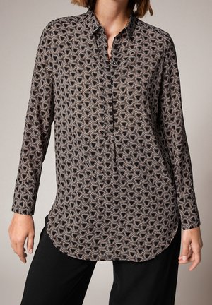 Kvinde iført en langærmet skjorte med sorte og beige geometriske hexagonmønstre, kombineret med sorte bukser.